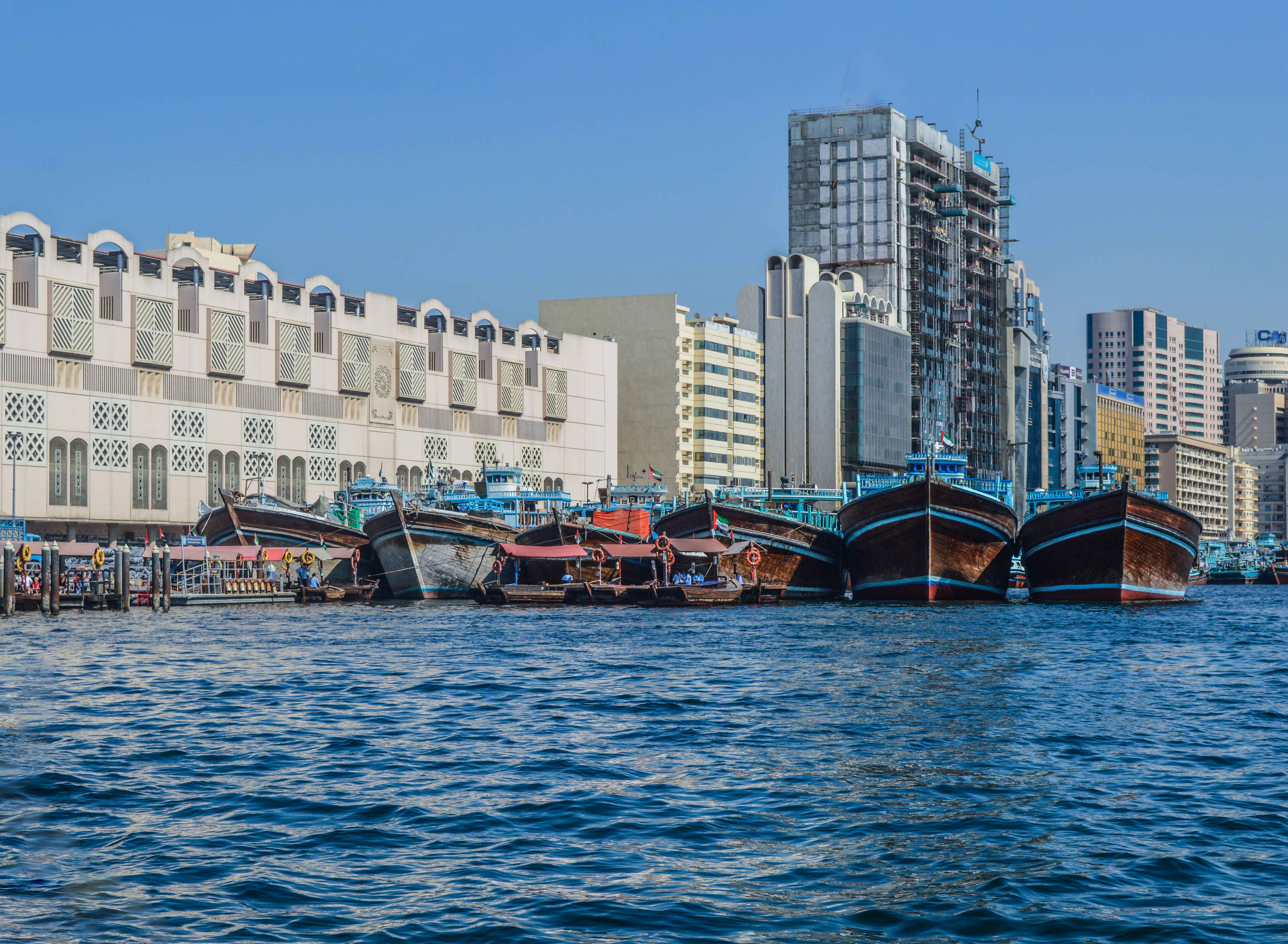 The image depicts a waterfront scene with traditional wooden boats docked along a pier. In the background, there is a mix of modern and traditional architecture, including a large, ornate building with intricate designs and several high-rise buildings under construction or renovation. The water is calm, and the sky is clear, suggesting a bright, sunny day.